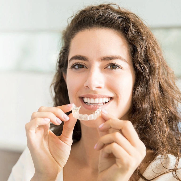Young woman holding clear orthodontic aligner, smiling in bright indoor setting.