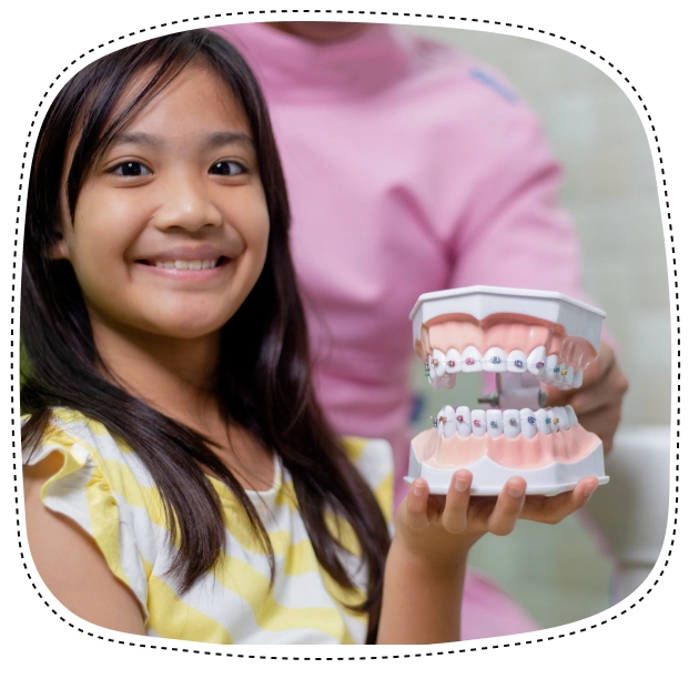 Young girl smiling while holding orthodontic braces model in dental office setting.