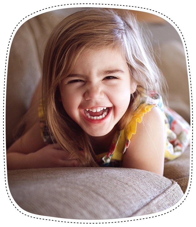 Smiling young girl with healthy teeth leaning on pillow in bright, warm indoor setting.