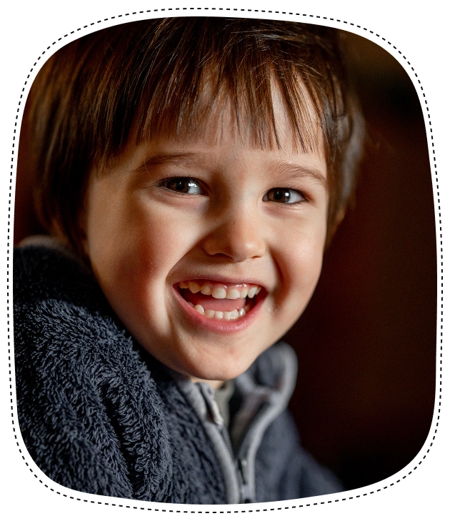 Young boy smiling brightly against dark background, wearing gray sweater.