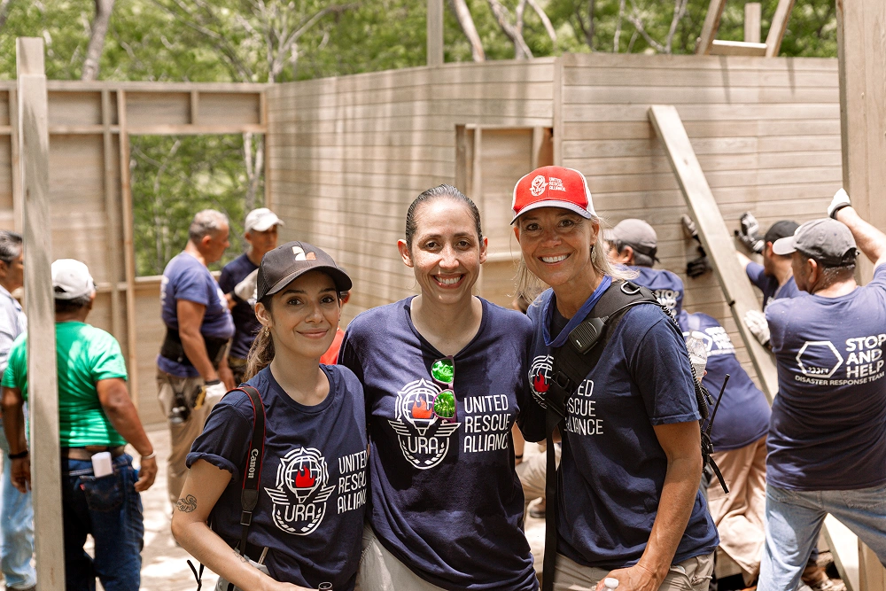 Three volunteers in matching shirts smile during outdoor community construction project.