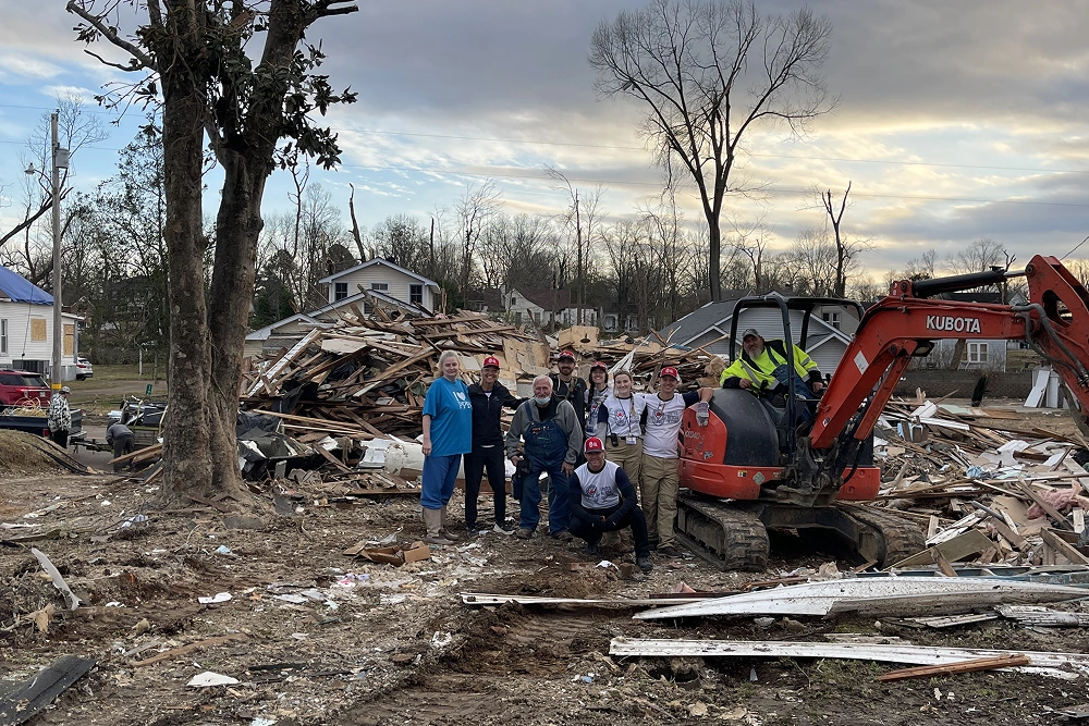 Group of people gathering at debris site with heavy machinery and bare trees visible.