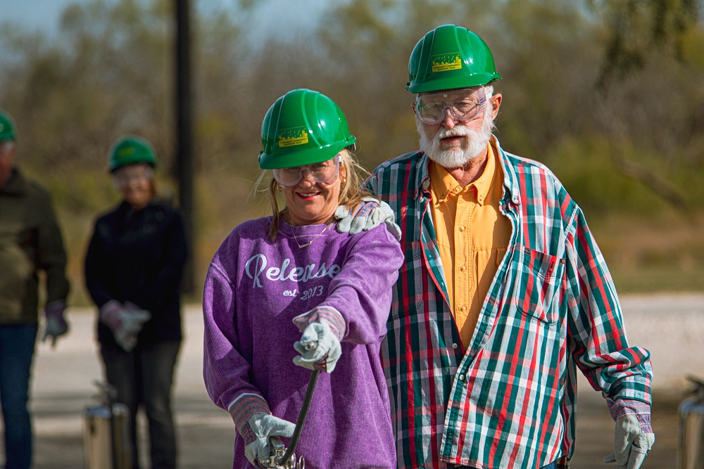 Two people wearing green hard hats and colorful shirts walk outdoors together during daytime.