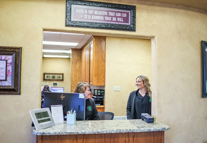 Two dental staff members stand behind reception desk in bright, welcoming clinic office.
