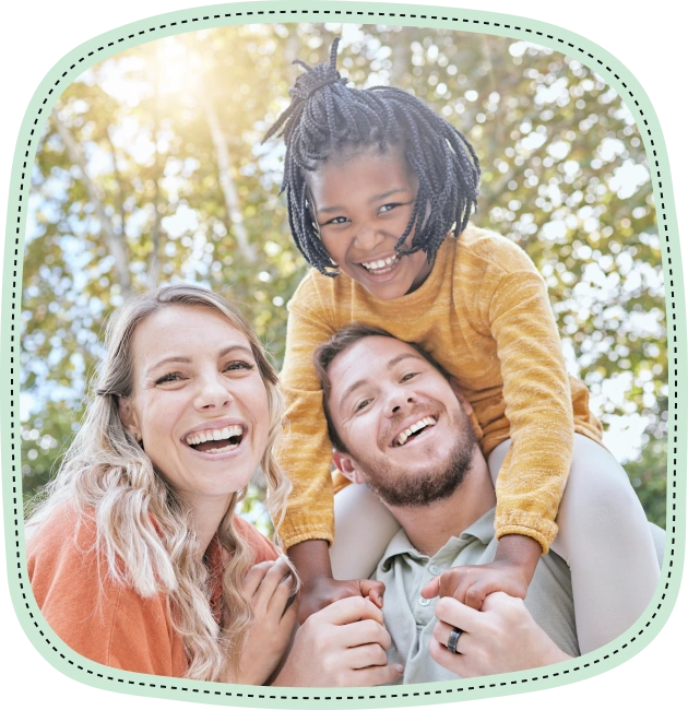 Happy family of four smiling outdoors in natural sunlight among trees.