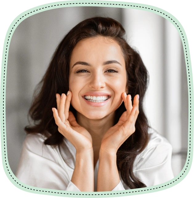 Happy woman in white shirt touching face, smiling at camera in bright clinical setting.