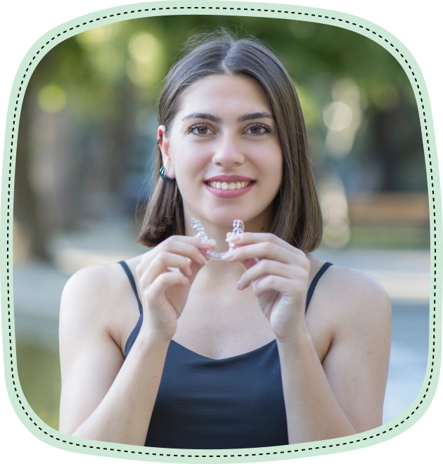 Young woman smiling outdoors, holding clear dental aligners in front of her face.