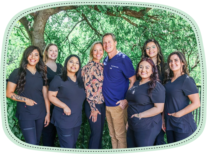 Professional dental team of eight smiling staff members posing together outdoors against green foliage.
