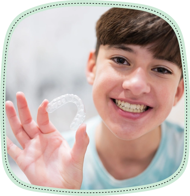 Smiling young boy holding clear dental aligners in front of his face indoors.