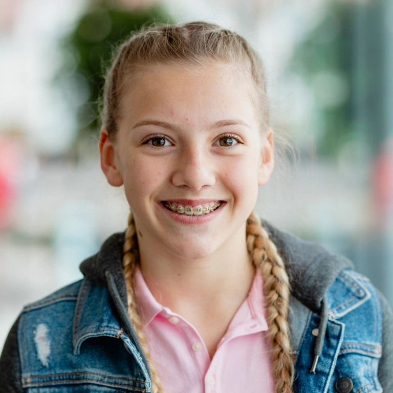 Smiling young girl with braided hair wearing denim jacket and pink shirt in bright indoor setting.