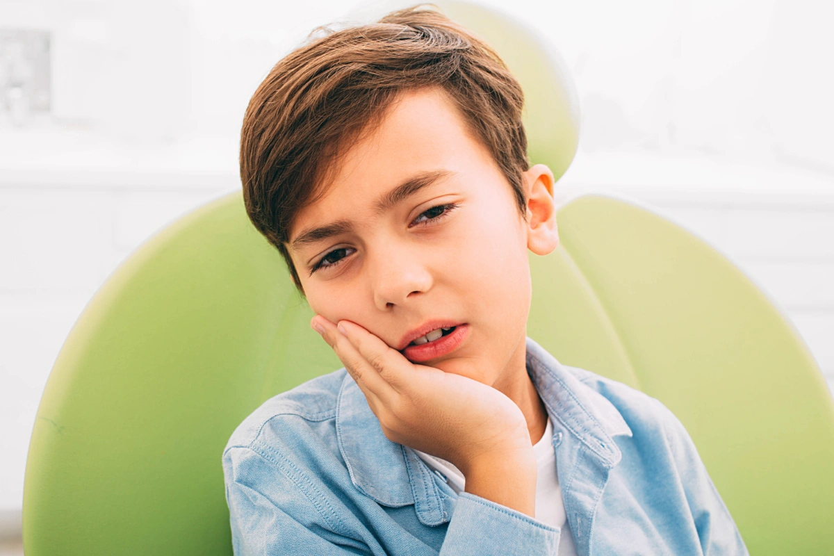 Young boy with hand on cheek against green background, appearing thoughtful or experiencing tooth discomfort.